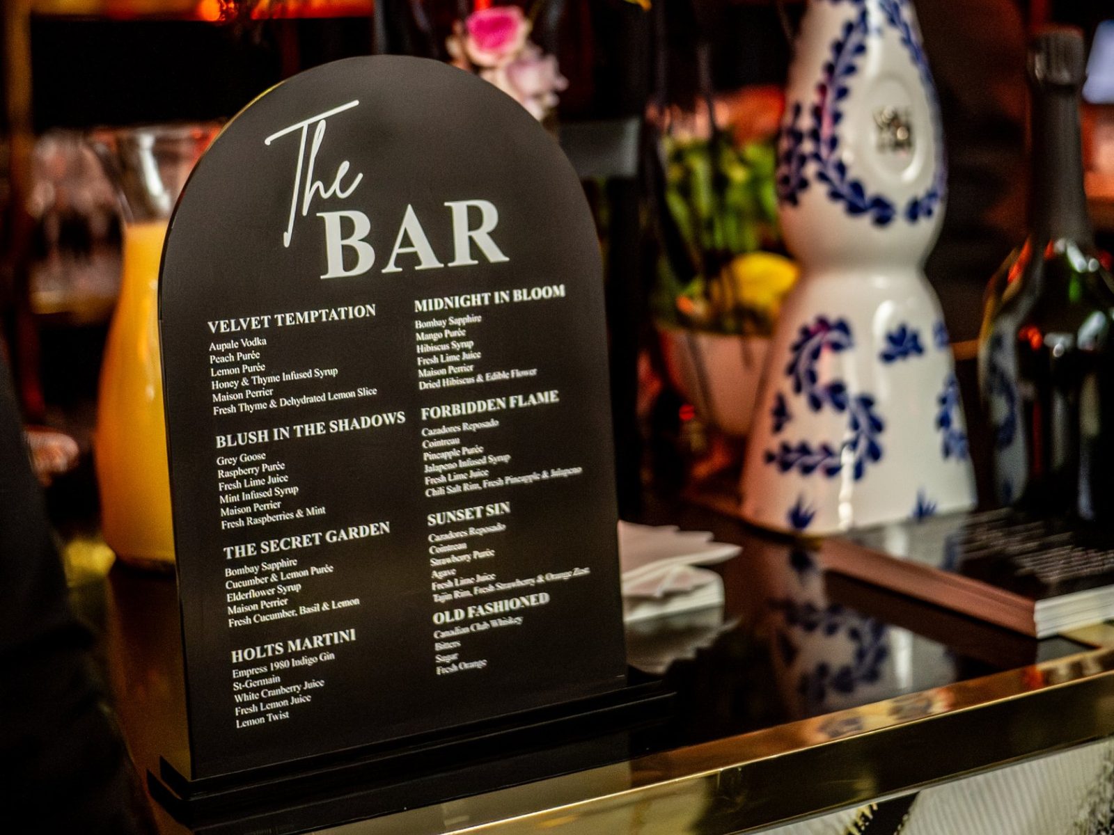 A signature cocktail menu on a brass bar framed by flowers